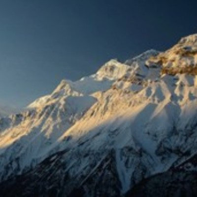 Photographed by Jonathan Duncan, snow-covered Annapurna Himalayan Range, illuminated by the golden light of a setting sun against a clear blue sky.