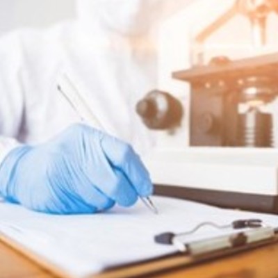 A lab worker wearing a protective and gloves takes notes on a clipboard in front of a microscope.