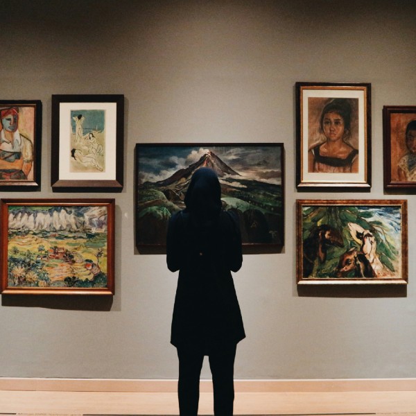 A person stands facing a wall of framed, fine art paintings.