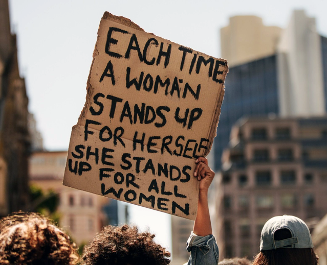 Protesting sign says "Each Time a Woman stands up for herself she stands for all women"