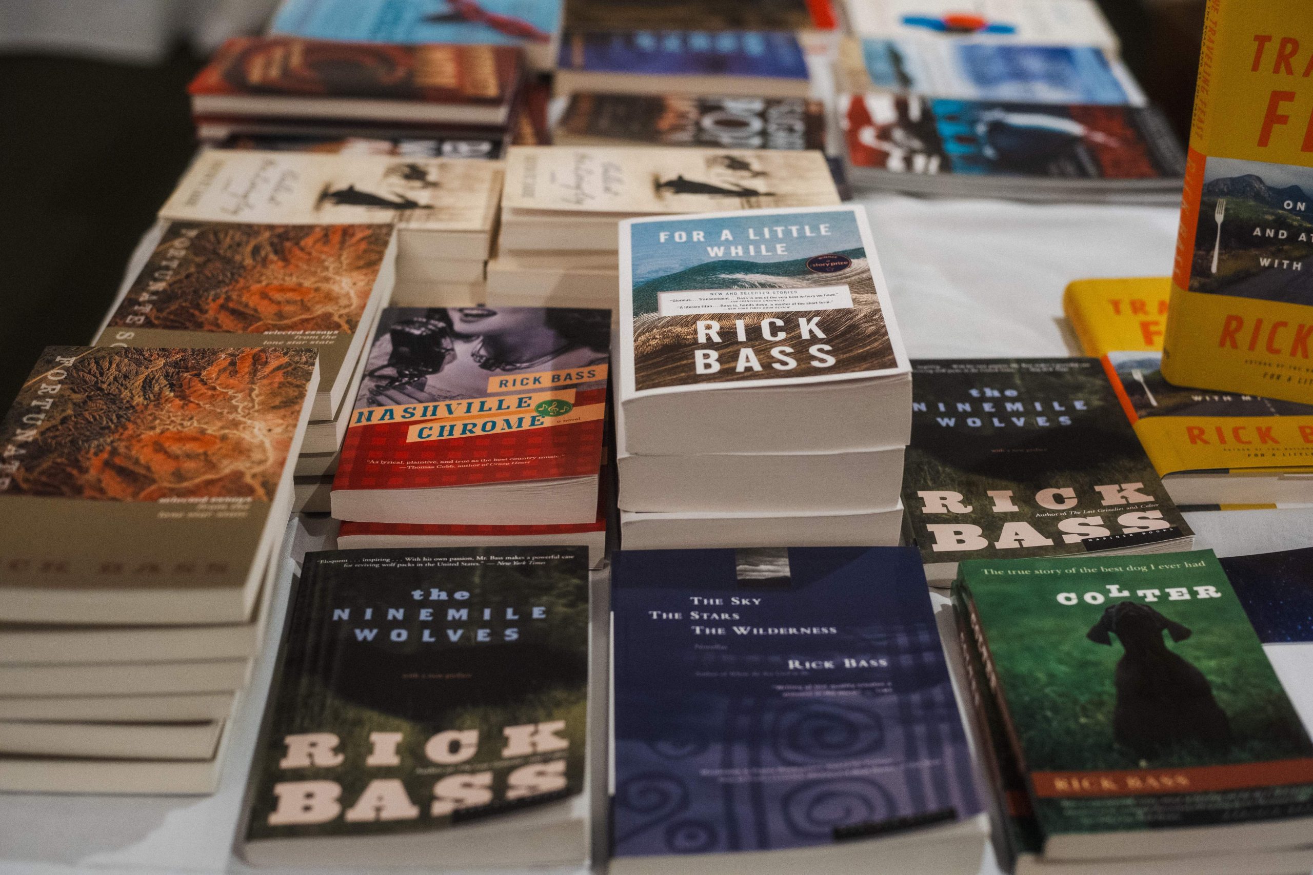 A table covered with stacked copies of books by author Rick Bass, including "The Ninemile Wolves," "Nashville Chrome," "For a Little While," "The Sky The Stars The Wilderness," and "Colter," displayed at a Stonecoast MFA event.