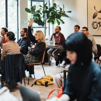 a woman with her hand up at a business meeting with other people, plants and paintings around her