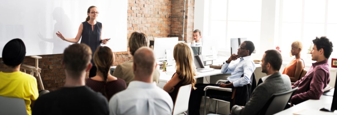 a group of people seated watching a young female instructor at a workplace training or business seminar