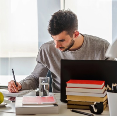 a man in a gray sweater writing on a pad, with a monitor and stack of books in the foreground