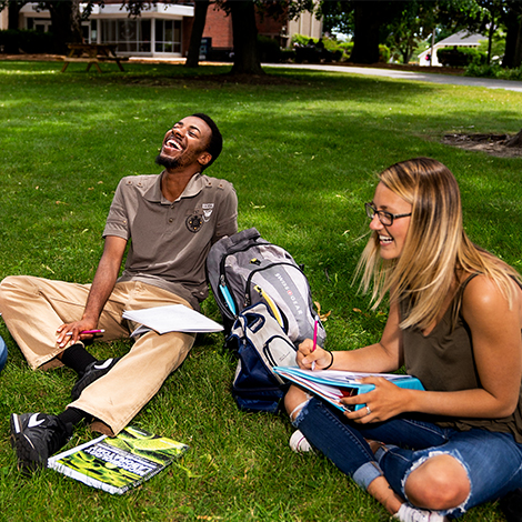Students sit in the grass, laughing, while taking notes in notebooks.