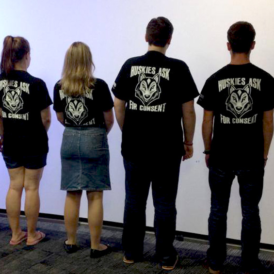 Four students face a projector screen to display the back of their t-shirts. Printed on the t-shirts are the words, ‘Huskies ask for consent’ and an illustration of a husky.