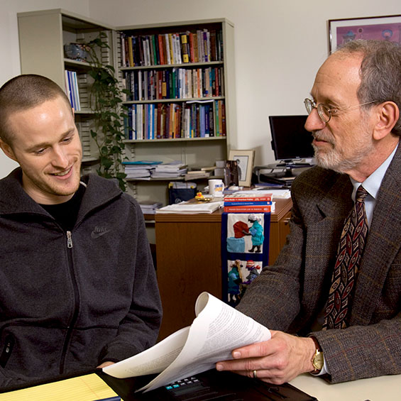 A student meeting with an advisor in their office. Metal bookshelves appear in the background.