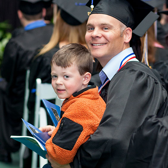 A graduating student smiles at the camera. Their child is seated on their lap and holds a commencement program. Other graduates appear in the background.