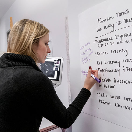 A person wearing a black sweater and writing on a piece of oversized paper taped to the wall.