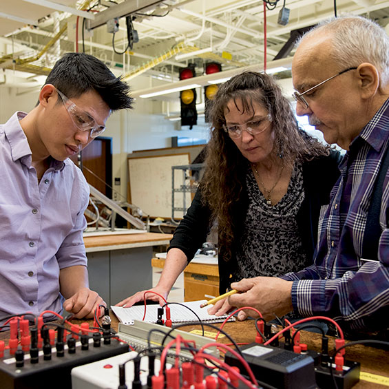 An engineering faculty member discusses an assignment with two students wearing safety glasses. Electrical equipment is clustered on the table.