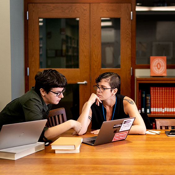 Two students wearing glasses working on a project in the Glickman Library on our Portland campus. On the table are a few books and two open laptop computers.