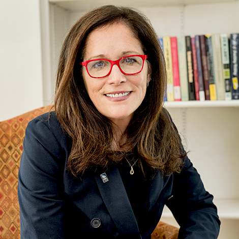 President Jackie Edmondson, wearing her signature red-framed glasses and a black blazer, is seated in an orange-patterned armchair next to a bookshelf.