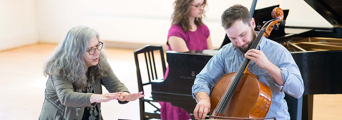 A faculty member conducts a student playing the cello in the foreground and a student playing the piano in the background during a rehearsal in Corthell Hall.