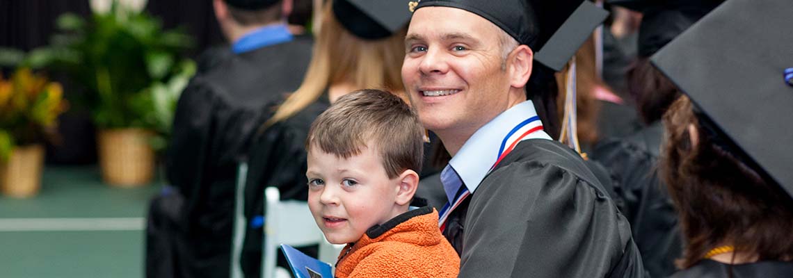 A graduating student smiles at the camera. Their child is seated on their lap and holds a commencement program. Other graduates appear in the background.