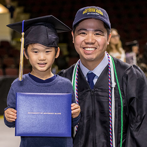 A graduate poses with their child at Commencement. The child is wearing the parent’s graduation cap and holding up their USM diploma cover.