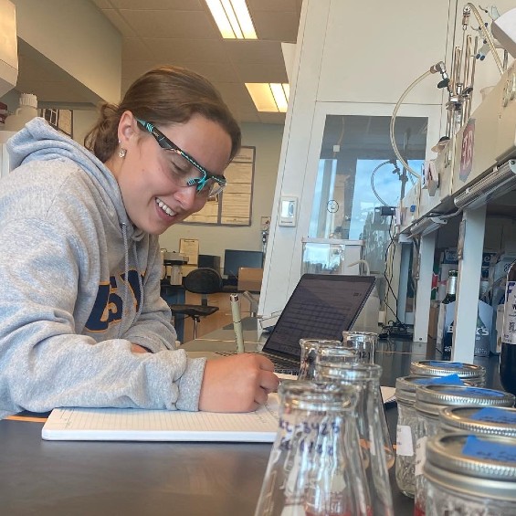 Student is sitting at a lab bench writing in her notebook. She is wearing safety glasses and there is a fume hood in the background