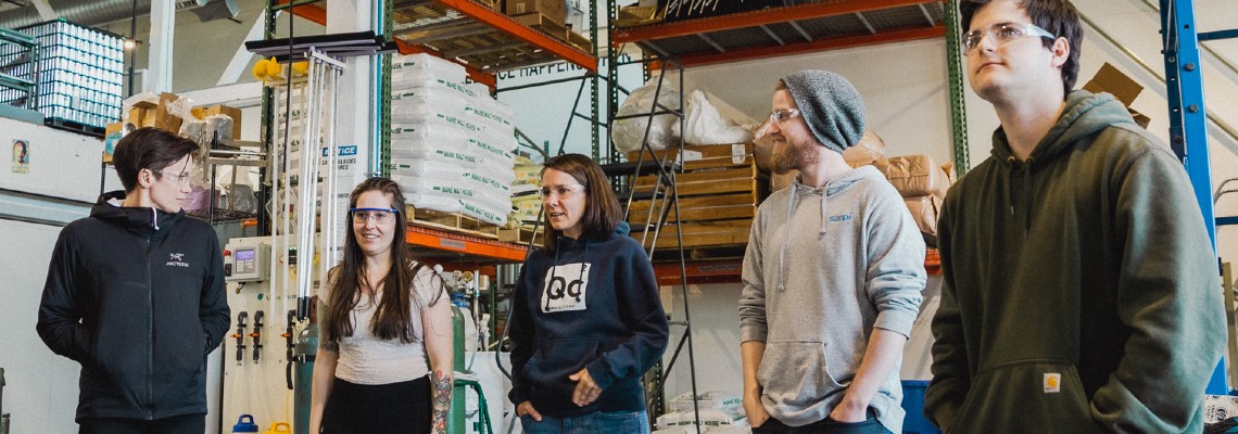 Five students are standing in a brewery production room with pallets of malt and materials behind them.