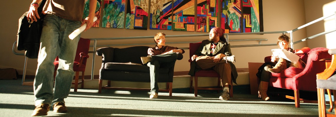 4 students. One is standing in the foreground and 3 are sitting in chairs/couches in the background. The closest student is casting a long shadow. There is a large modern art installation on the wall behind them.