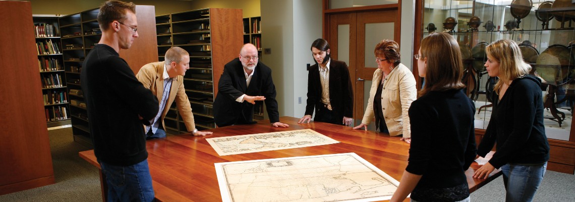 Seven people standing in a library positioned around a wood table while looking at two old maps.