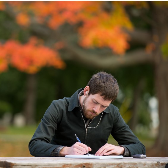 A student listening to headphones is writing in a notebook while seated outside in an out of focus fall scene.