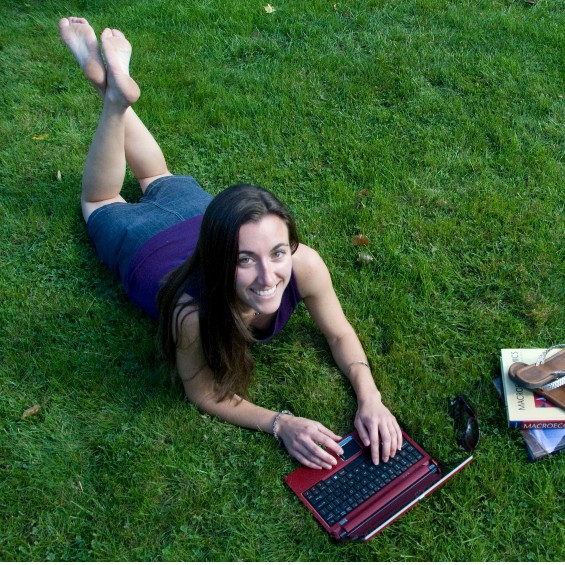 A barefoot student lays on freshly mowed grass and studies with a red laptop. Her sandals are stacked on top of her macroeconomics book.