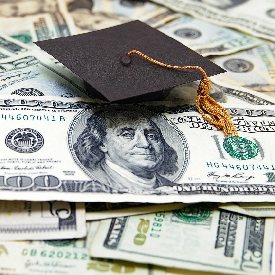 A black Mortarboard hat (graduation cap) with a gold tassel on a pile of American dollar bills. The 100 dollar bill is most prominent, but there are multiple 20 dollar bills and a 5 dollar bill visable.