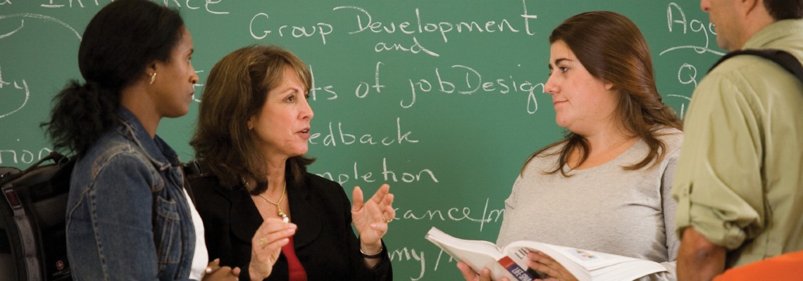 3 students and an instructor discuss while standing n front of a wall-length chalk board. "Group Development and Elements of Job Design" is written on the chalkboard.