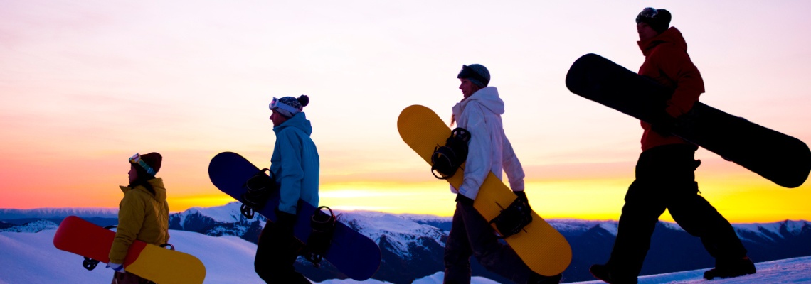 Four people with snowboards outside as the sun sets. There is snow on the ground, everyone is bundled up in jackets and hats, and the sky is clear.