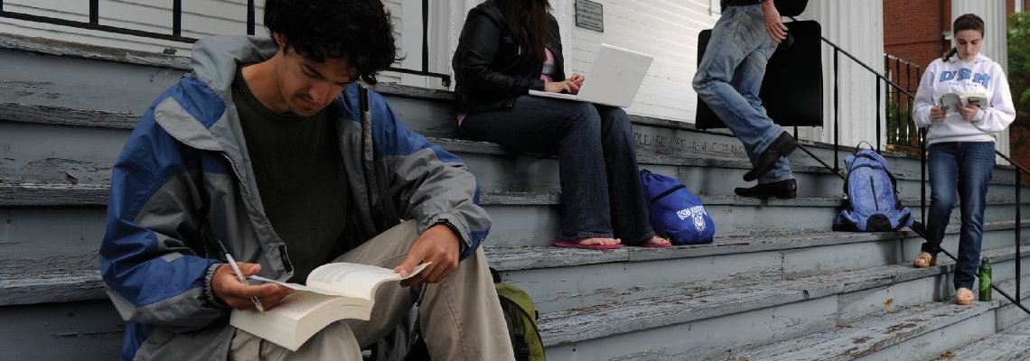 Students study on the steps of a building. The closest to the camera is pouring over a book, while a student further back in the perspective is working on a laptop.