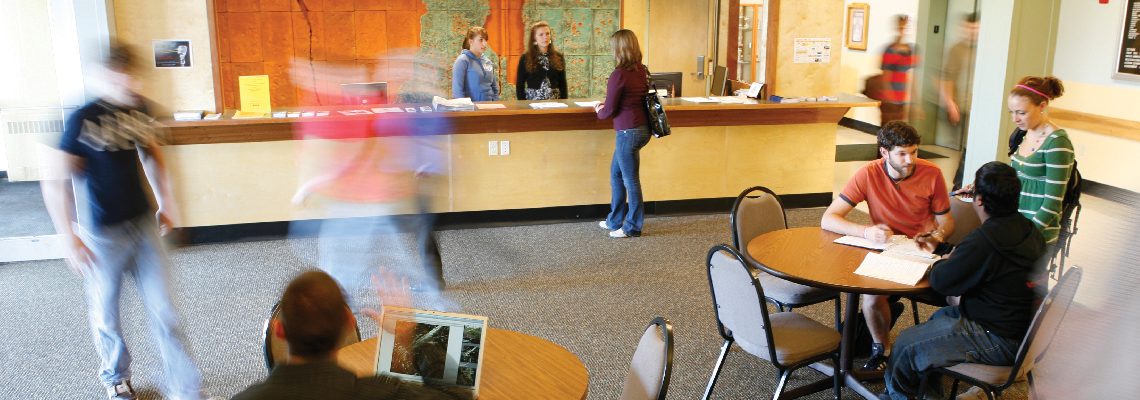 A bright image of students at desks in the foreground with a student consulting some people behind a help station. The photo has a long exposure and it has captured a blur where a student was walking quickly.