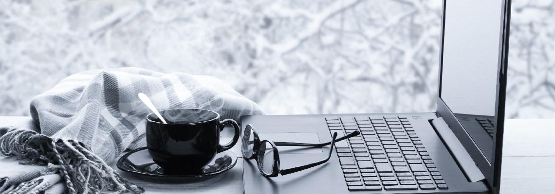 A wintery scene with brush covered with snow in the background. In the foreground, rests a shiny slim black laptop with a pair of black glasses resting on the mousepad. A steaming hot drink and a black, grey and white scarf rest to the left of the laptop.