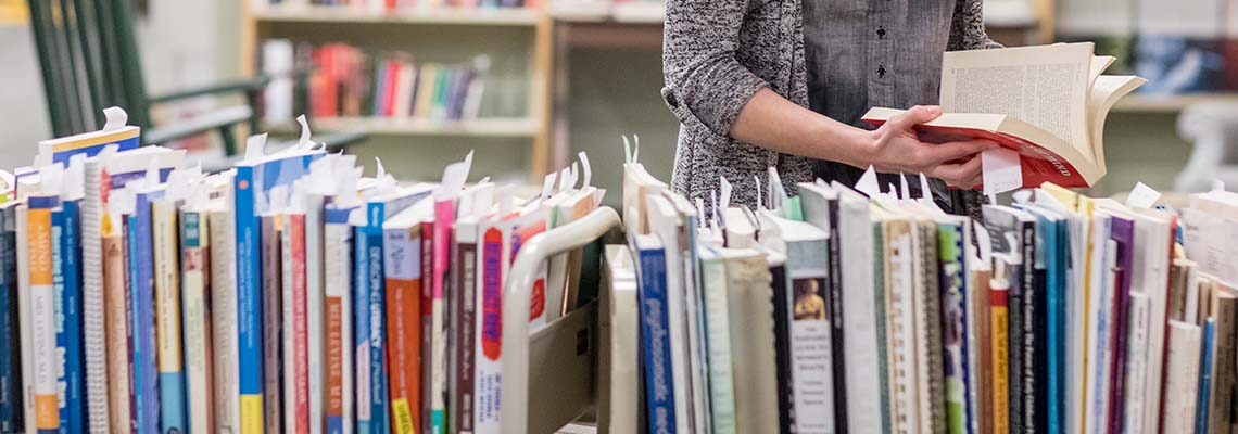 A student browsing books lined up neatly on metal book carts in our Lewiston library. Small slips of paper stick out of each book.