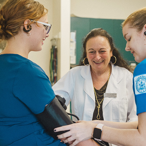 A nursing faculty member smiles as two undergraduate nursing students practice using a blood pressure cuff.