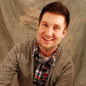 Brandon is Wearing a multi-colored plaid shirt with a gray cardigan. There is a tan backdrop behind him and he is smiling.