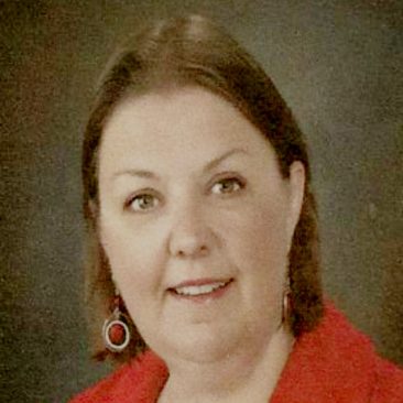 Deb Gillespie has white skin and light brown hair and brown eyes. She is wearing a red blazer.