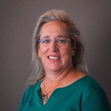 A headshot of Leslie. She has glasses, long hair, a green top, and is smiling.