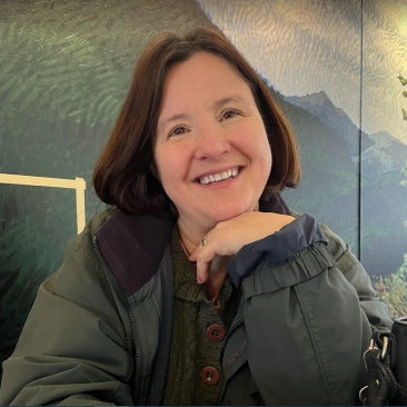 This is a photo of Katharine Thayer. She is wearing a green jacket, has short brown hair, and is smiling.