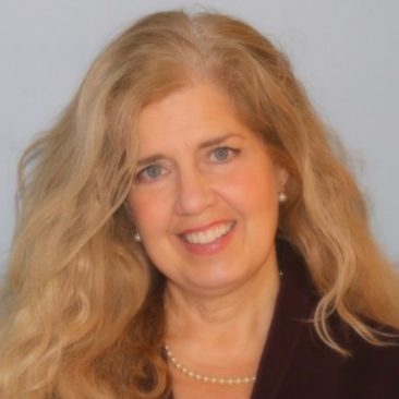 Photo of Suzanne Parkman, who is wearing a burgundy suit, and has blond hair, light skin, and blue eyes. The photo is a head and shoulder shot with a white background and she is smiling.
