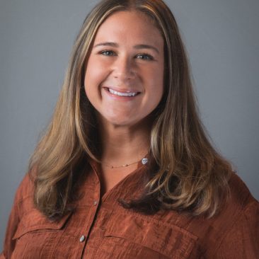 Suzanne has long brown hair and is smiling and is wearing a brown shirt.