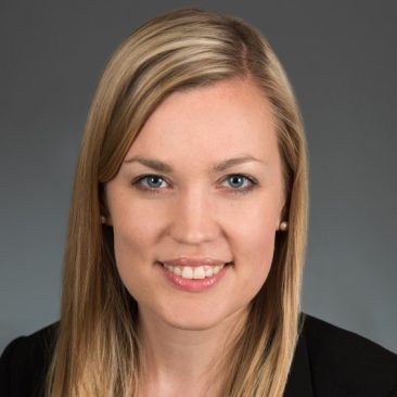 Photo of Sarah Parker, who is wearing a blue top and black blazer, and has blonde hair, light skin, and blue eyes. She is pictured against a gray neutral backdrop and is smiling.