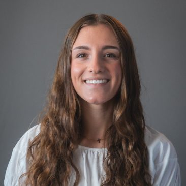 A smiling woman with long wavy brown hair and a white top in front of a gray background