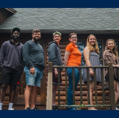 5 people standing in a row, in front of a log cabin