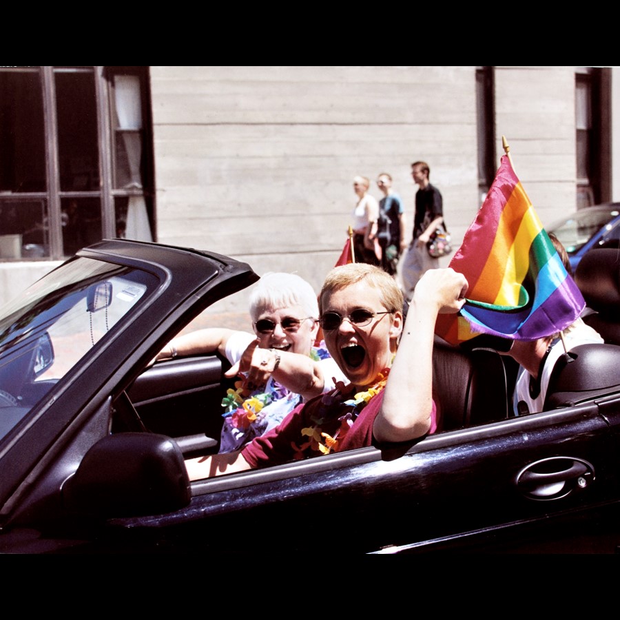 Two people sitting in a car reaching back holding on to a rainbow flag. 