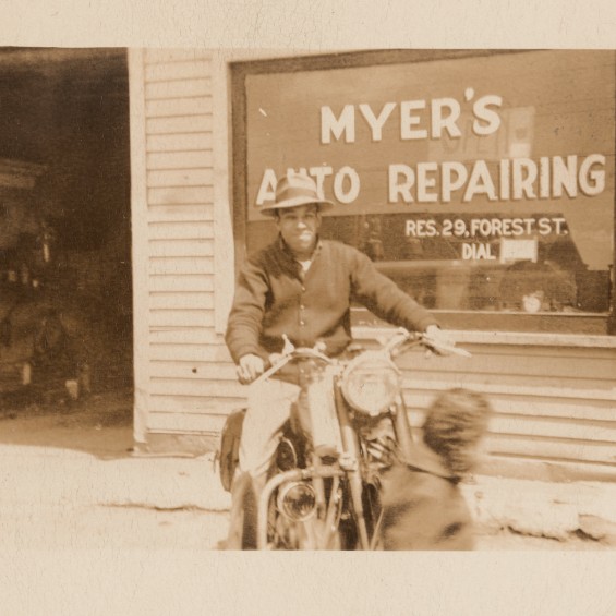 Photograph of a man on a motorcycle in front of a shop