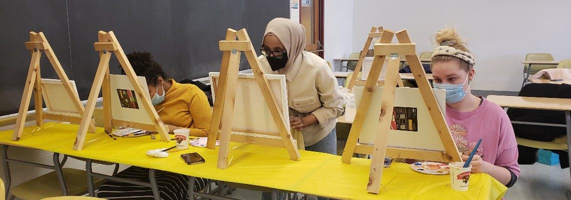 Three students sitting side by side, painting on art easels