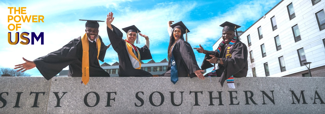 photo of 4 USM grads in cap and gown on the wall in front of the Bean Green