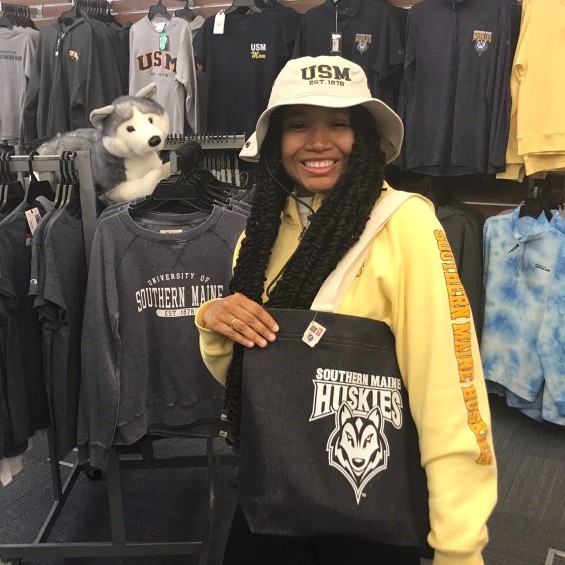 A student worker posing with University Store merchandise she is wearing a USM hat, sweater and is sporting a USM Huskies bag