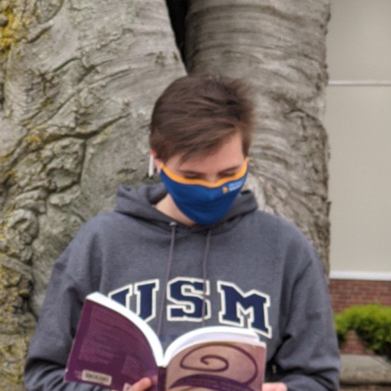 A student in a USM sweater reading a textbook in front of a tree on the Portland Campus