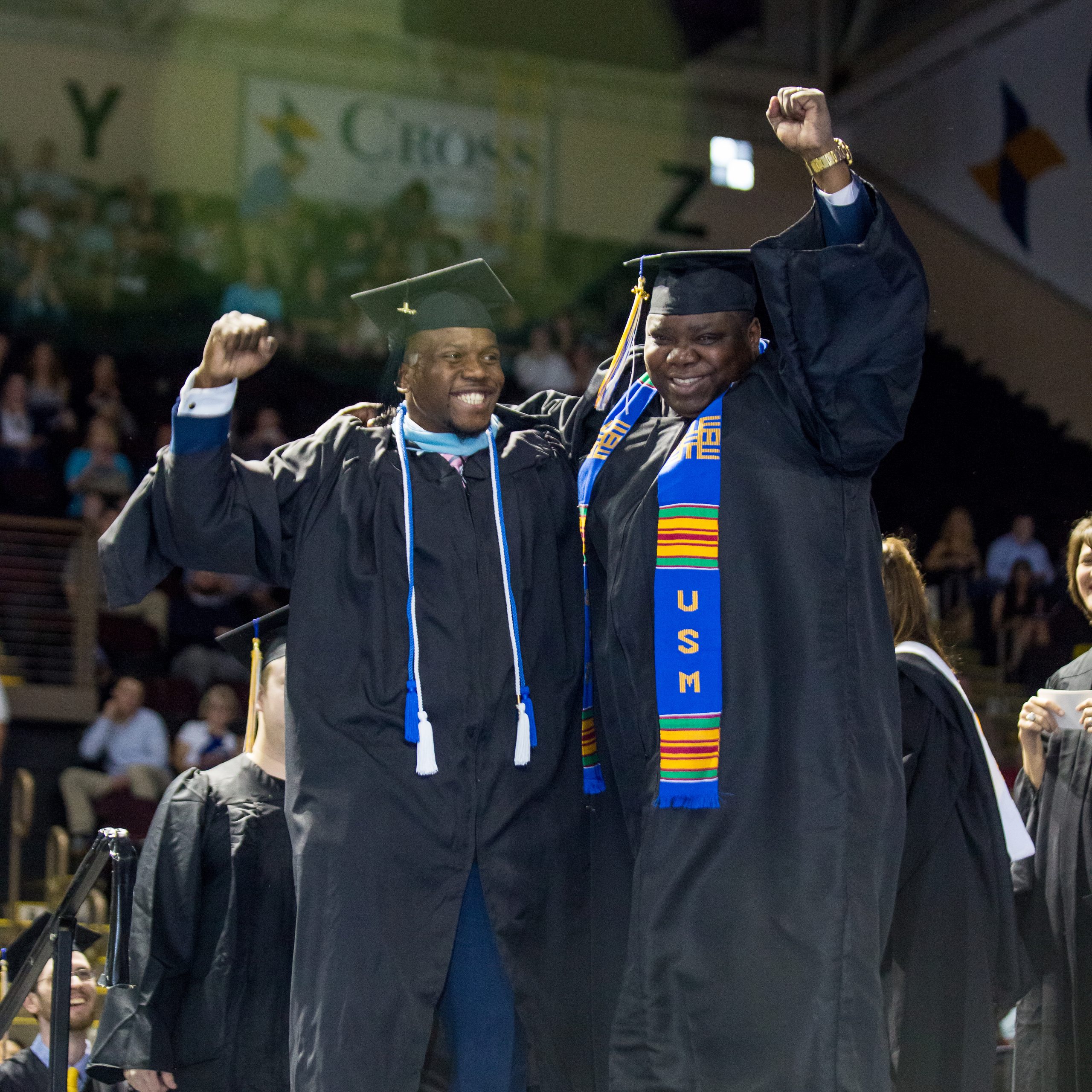 Two graduates in regalia, crossing the stage with broad smiles and fists raised in the air.