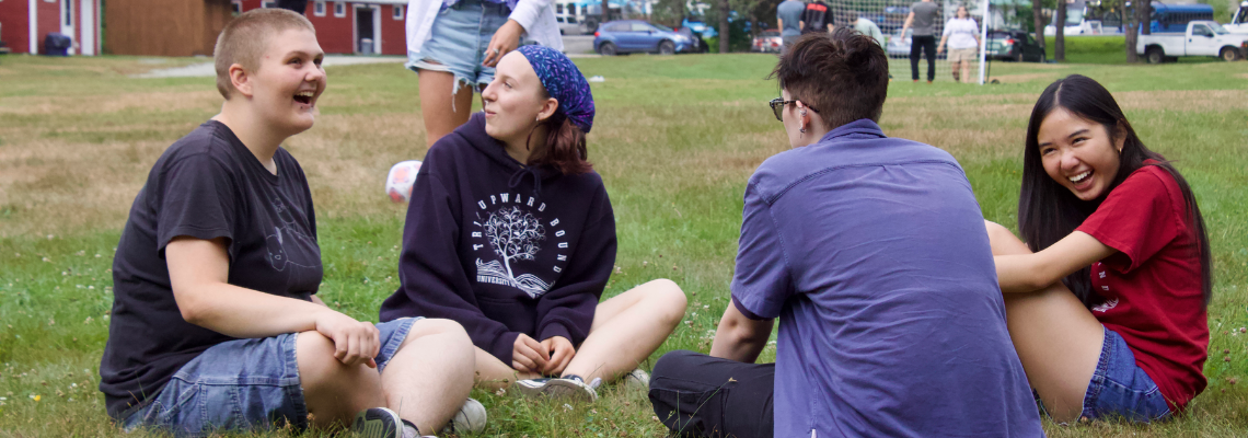 Four students sit outside in the grass laughing